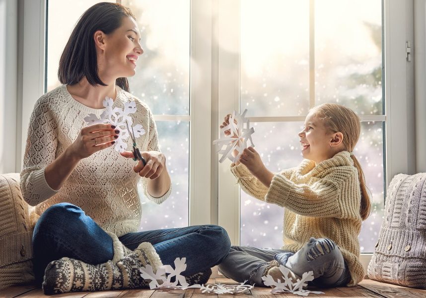 Merry Christmas and happy holidays! Happy loving family sitting by the window and making paper snowflakes for decoration windows. Mother and child creating decorations.
