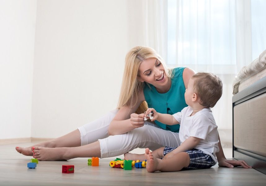 Beautiful mom is spending time with her baby in bedroom. They play with toys. The parent and her chills are looking at each other and smiling